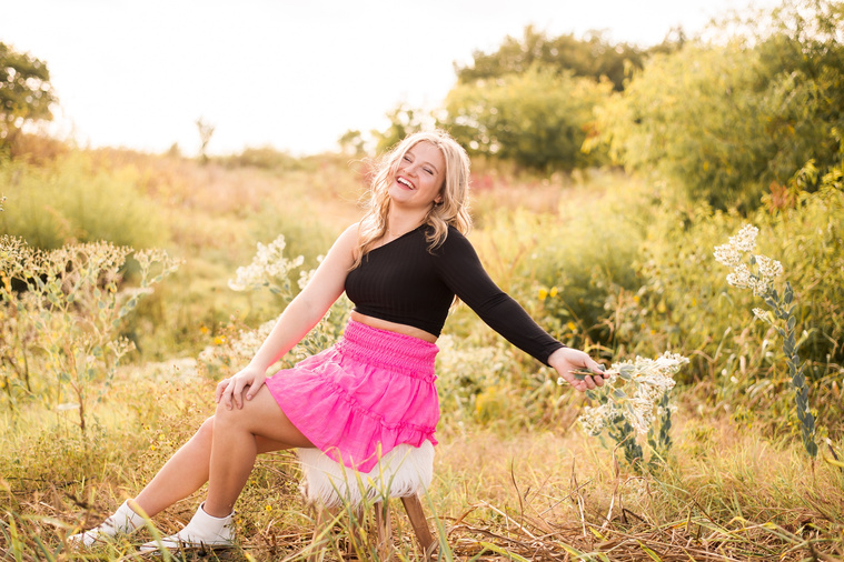 high school senior girl with blond hair wearing a bright pink skirt and black shirt, sits in a golden field in Oklahoma holding a flower laughing for her photo session