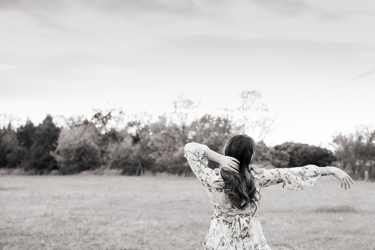 black and white photo of a high school senior girl dressed in boho style standing in a field with trees and a sunset behind her with one arm out in a carefree way in Oklahoma City, Oklahoma