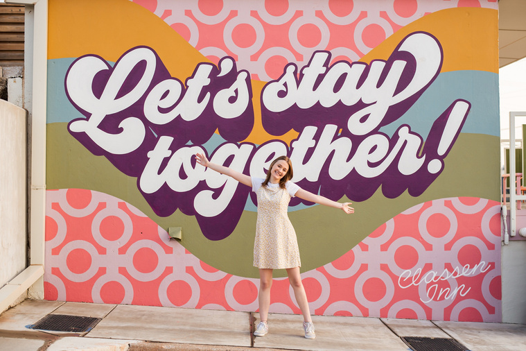 high school senior girl stands in front of a colorful wall mural that says Let's stay together on the wall of the classen inn in midtown Oklahoma City with her arms open and smiling
