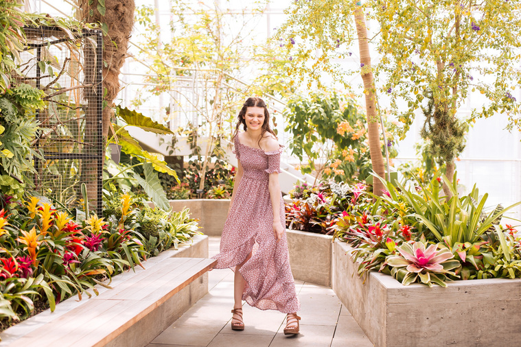 high school senior girl with long brown hair in a red and white dress stands mid twirl and smiles while she holds her skirt on a path with tropical plants all around inside the Crystal Bridge at Myriad Gardens in Oklahoma City 