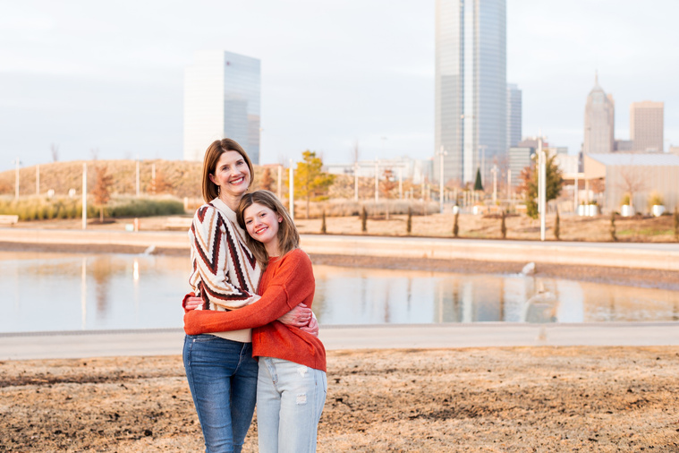 a mom and her teen daughter hug each other at Scissortail Park with the Oklahoma City skyline behind them
