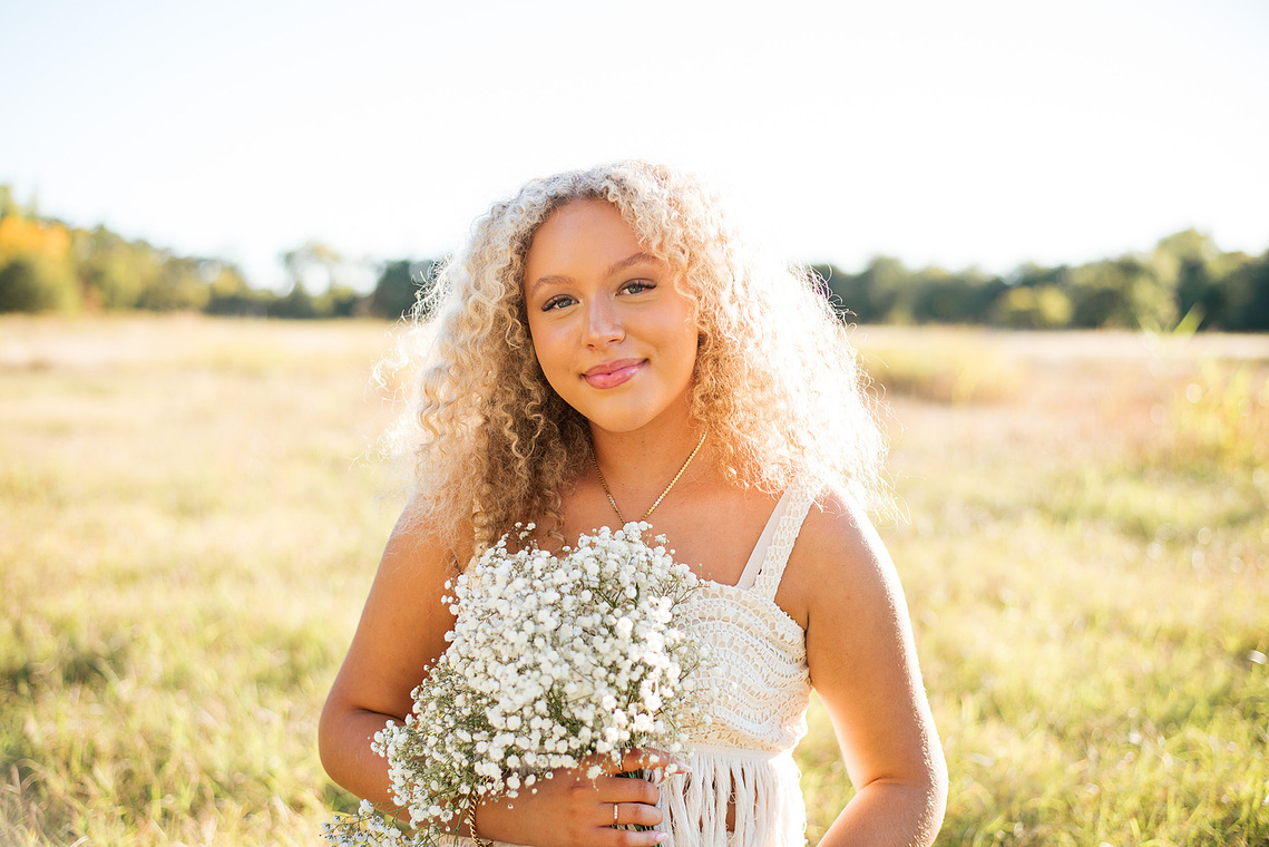 fall photo of a beautiful senior girl with curly blond hair wearing a white top and holding a flower bouquet in the middle of a field at Martin Nature Park in Oklahoma City
