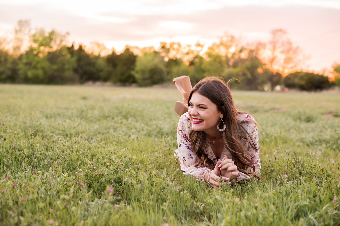 high school senior girl dressed in boho style lays on her stomach and smiles to the side  in a field with trees and a sunset behind her in Oklahoma City, Oklahoma
