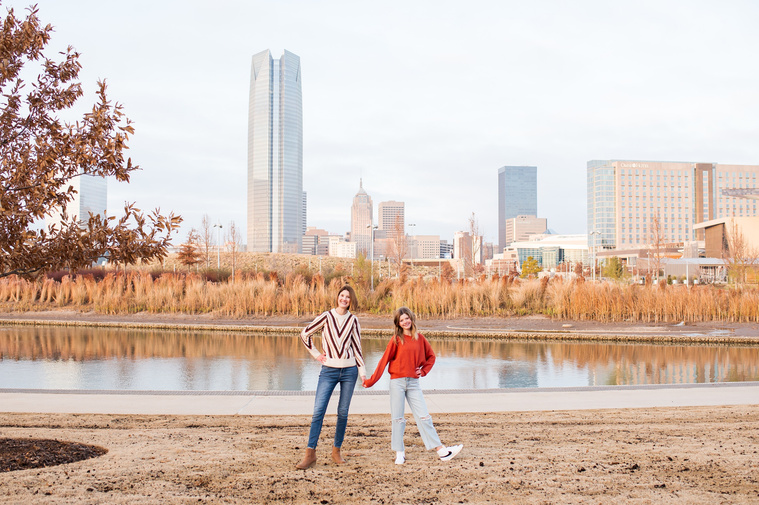 a mom and her teen daughter hold hands and kick out their heels at Scissortail Park with the Oklahoma City skyline behind them