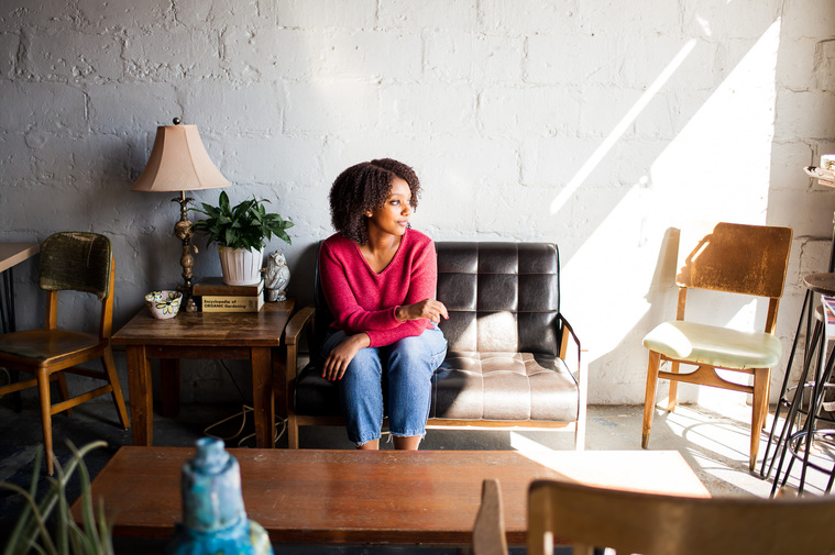 wide angle shot of a young black girl wearing a red top and jeans sitting on a couch in a coffee shop looking to the side with a coffee table and chairs around her