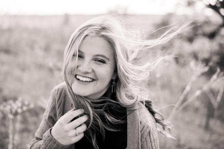 black and white high school senior portrait of a girl sitting in a field smiling with the wind blowing her hair and holding the ends of her hair with one hand