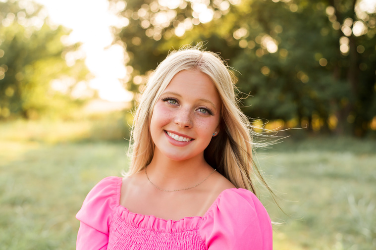 Blond high school senior girl in a bright pink shirt smiles for a photo in a field with trees and sunlight behind her in Oklahoma