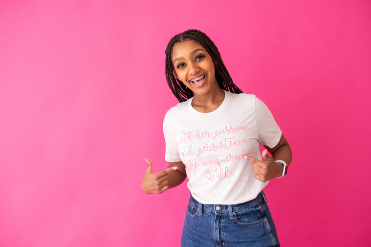 young black girl with long braids stands in front of a hot pink background wearing a t-shirt and jeans and pointing to the words on the shirt while smiling 