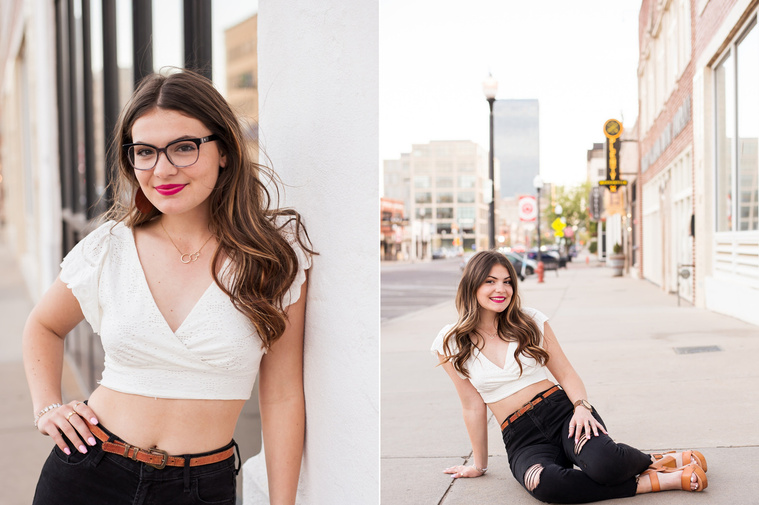 two photos of a high school senior girl with long brown hair in a white top and black jeans, one with glasses, standing and sitting on the sidewalk around buildings in Automobile Alley in Oklahoma City, Oklahoma