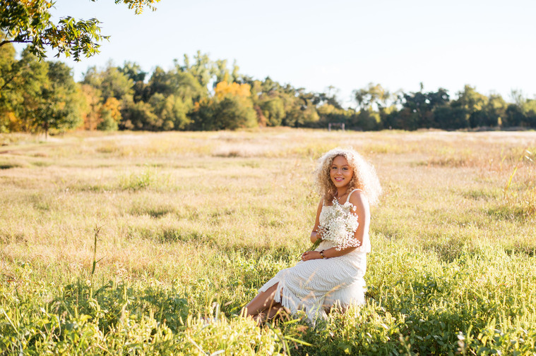 high school senior girl with blond curly hair wearing a white dress holding a bouquet of flowers sits in an open green field with bright sunlight surrounding her at Martin Nature Park in Oklahoma City