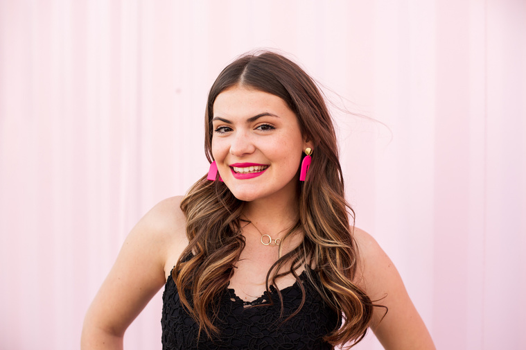 high school senior girl with long curly brown hair stands in front of a pink wall in Oklahoma City wearing a black top and pink earrings smiling at the camera