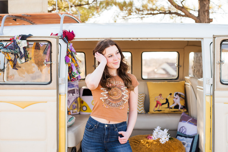 senior girl with brown hair wearing a graphic t shirt and blue jeans stands in front of an open VW bus with retro decor with a hand in her hair smiling