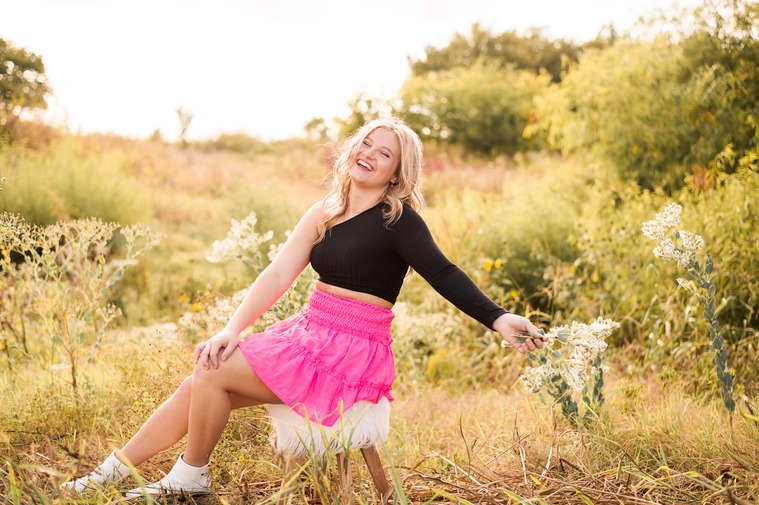 high school senior girl wearing a black top and pink skirt sits on a boho stool holding flowers and laughing with grass and plants behind her in a field near Tuttle Oklahoma
