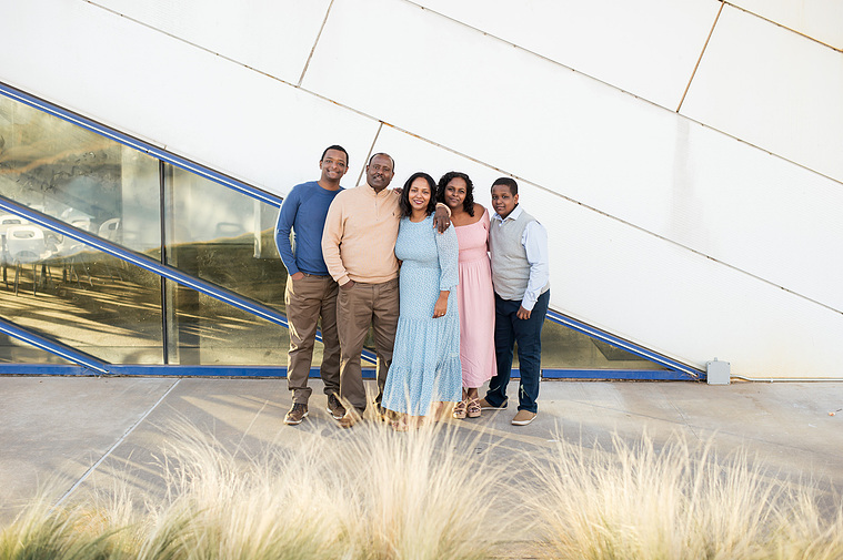 parents hug and smile together with their 3 children dressed in soft colors in front of a the angled lines of a boathouse near the Oklahoma River