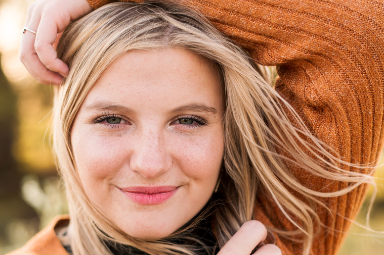 close up photo of a high school senior girl with blond hair wearing a rust colored sweater with her arm framing her face in a golden field in Oklahoma 