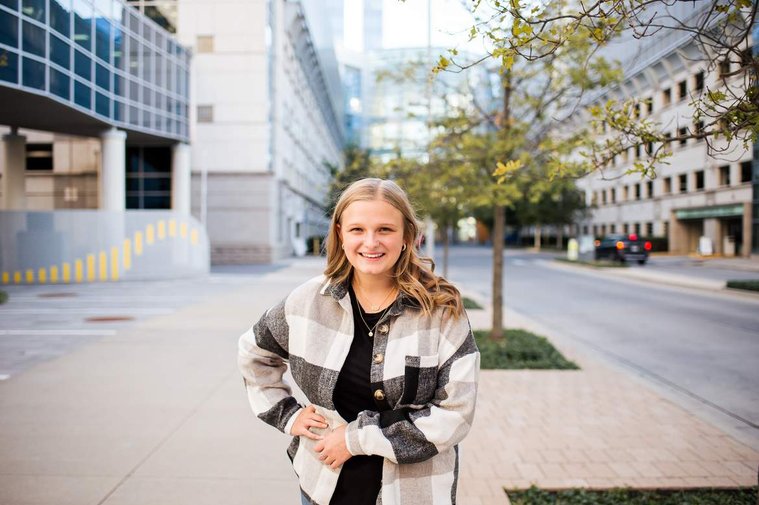 high school senior girl stands and smiles with her hands on her waist in a black and white jacket in front of the Devon buildings in Oklahoma City