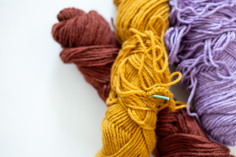 close up brand photo of a brown, yellow and purple roll of yarn stacked together with a green crochet needle sticking out of yarn with white space on the left