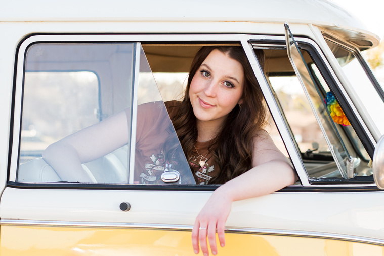 high school senior girl sitting in the front passenger side of a yellow and white VW bus with her arm draped out the window and her head at the window smiling
