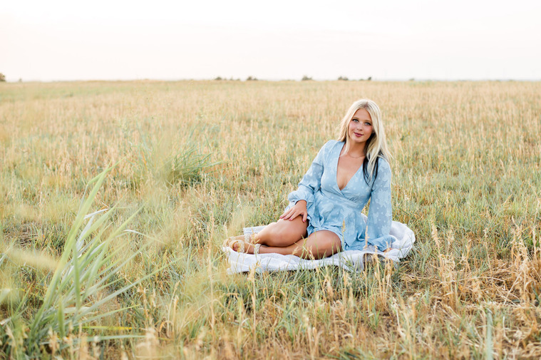 high school senior girl with blond hair and blue dress sits on a blanket in the middle of a rustic country field with grass in foreground in Tuttle Oklahoma
