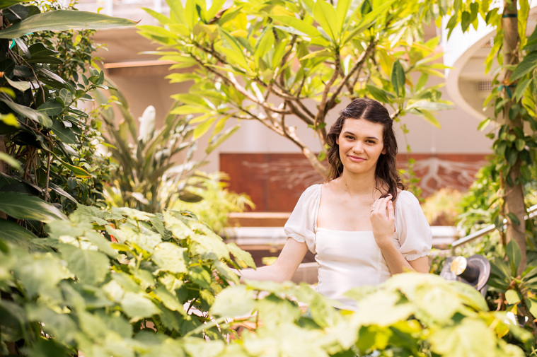 high school senior girl with brown hair in a white blouse sits and smiles softly among tropical plants and leaves all around her at the Crystal Bridge at Myriad Botanical Gardens in Oklahoma City