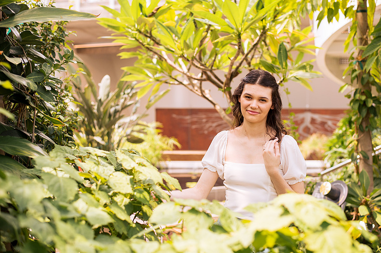 high school senior girl with brown hair in a white blouse sits and smiles softly among tropical plants and leaves all around her at the Crystal Bridge at Myriad Botanical Gardens in Oklahoma City