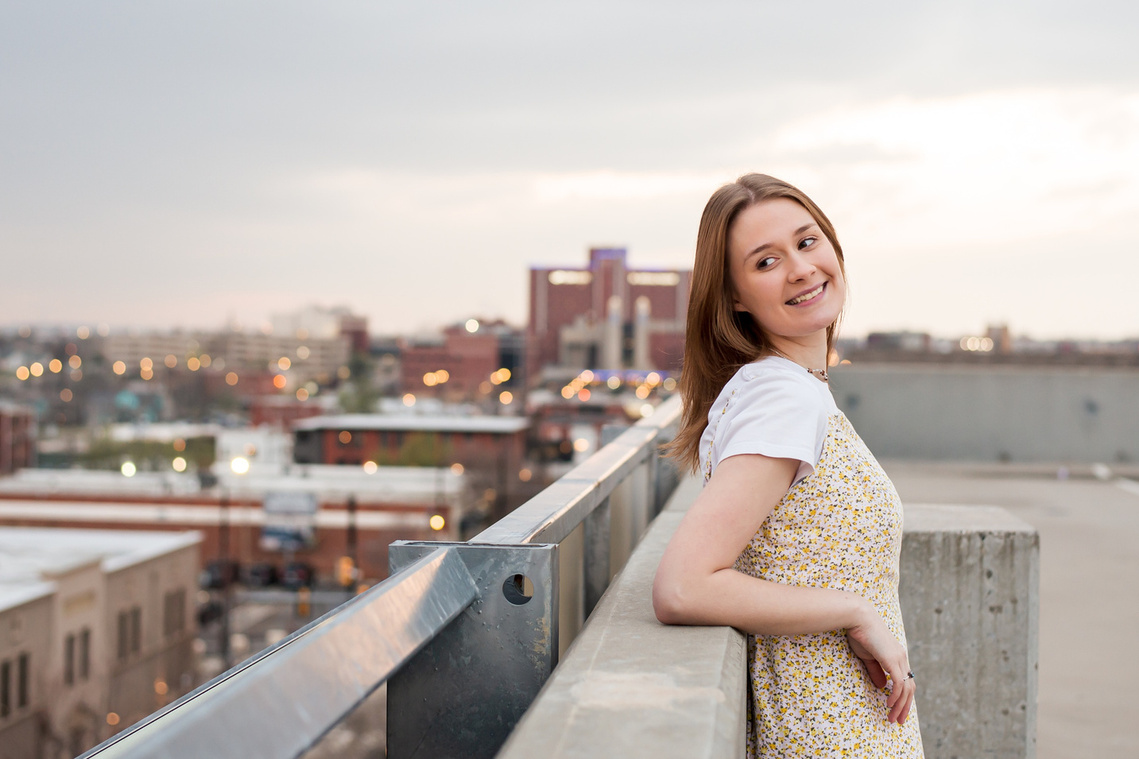 high school senior girl leans against wall on the rooftop of a parking garage in midtown Oklahoma City and smiles over her shoulder