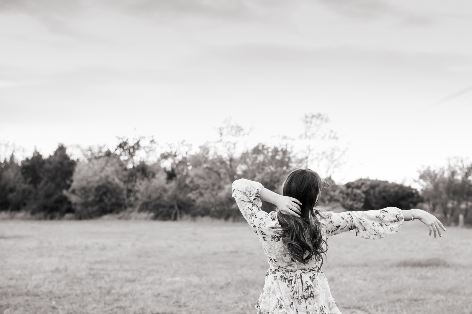 black and white landscape photo of a senior girl facing away with one arm extended out and one on her head with a tree line behind her in a field in southwest Oklahoma City