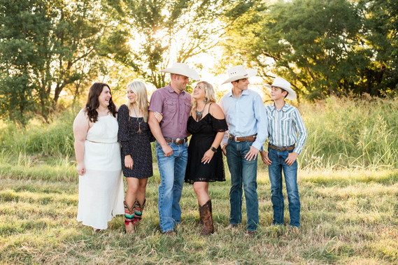 mom and dad with their 4 teen kids dressed in western outfits stand together and look at one another in a field with trees and grass around them in Oklahoma