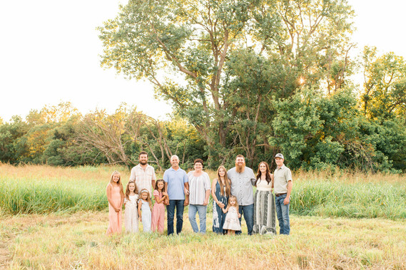 a grandma and grandpa stand with their adult children and families around them smiling together in  front of a tree line and tall grass in a field on a farm in central Oklahoma
