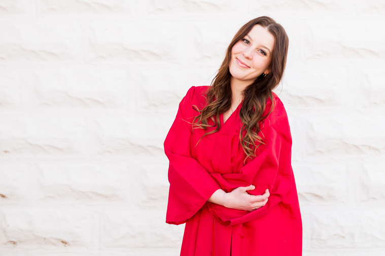 high school senior girl wearing her red graduation gown hugs her elbows and smiles in front of a white wall in Tuttle Oklahoma