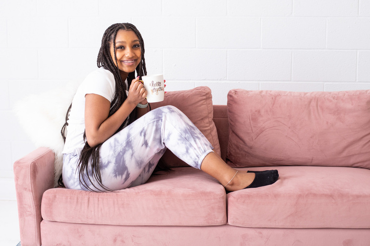 young girl with long hair sits cozily on a pink couch holding her mug and smiling for a product branding photoshoot in an Oklahoma City studio