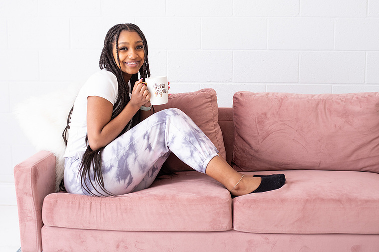 young girl with long hair sits cozily on a pink couch holding her mug and smiling for a product branding photoshoot in an Oklahoma City studio