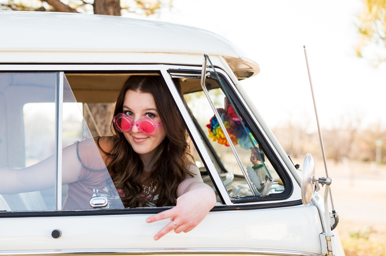 high school senior girl with brown hair wearing a t-shirt and bell bottoms sits in the passenger seat of a VW bus in a yard in Tuttle Oklahoma with the window open smiling with sunglasses and a peace sign