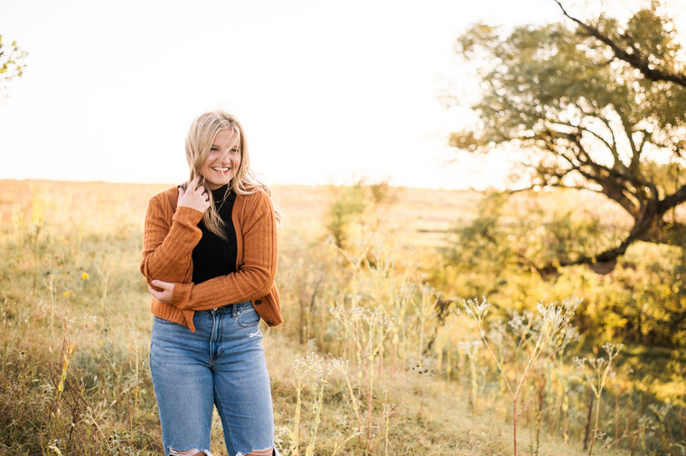 senior girl with blond hair wearing an orange sweater, black turtleneck and jeans stands in a field smiling in the fall with grass and trees behind her with hair blowing in her face
