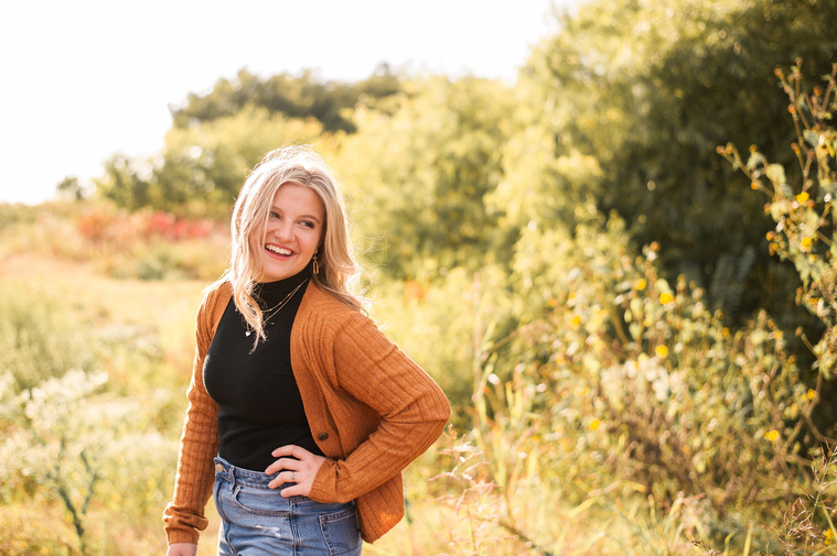 high school senior girl with blond hair wearing a rust colored sweater and jeans, stands in a golden field in Oklahoma looking over her shoulder and smiling for her photo session