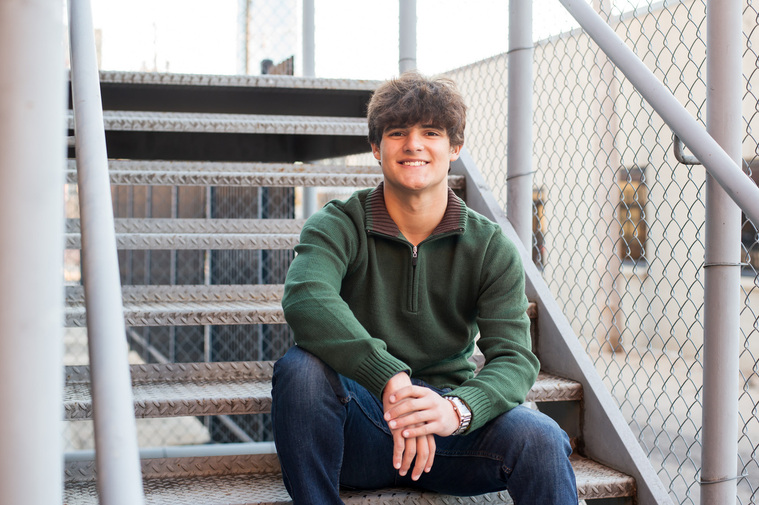 cute high school senior boy sits on metal steps in an alley in midtown OKC wearing a green sweater and jeans and smiling