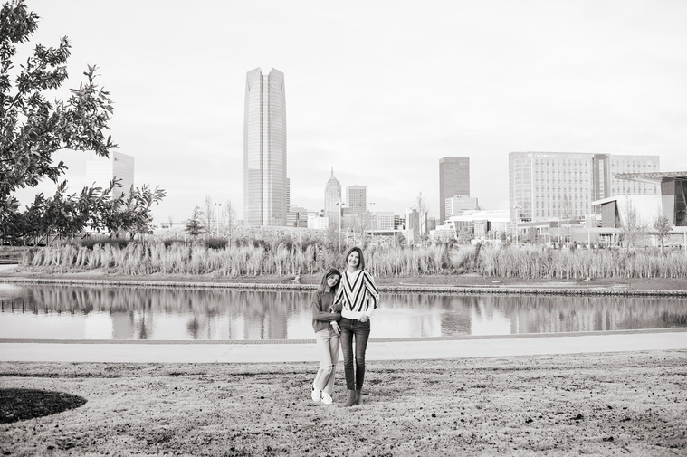 a mom and her teen daughter hold hands and stand together at Scissortail Park with the Oklahoma City skyline behind them