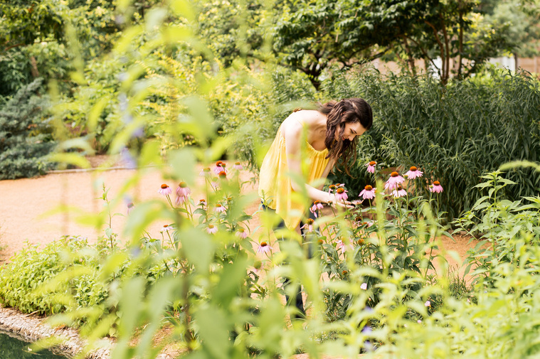high school senior girl with long brown hair wearing a yellow top and jeans stands among flowers and plants admiring them at Myriad Gardens in Oklahoma City