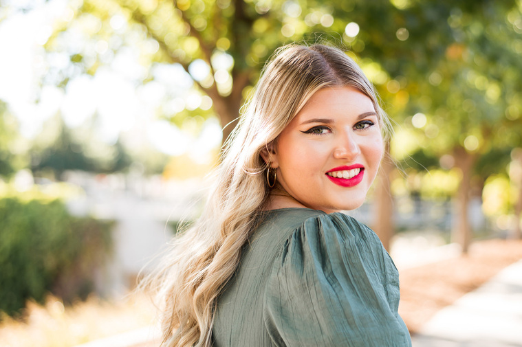 high school senior girl with long blond hair wearing a green dress looks over her shoulder and smiles, standing on a sidewalk with green trees and sunlight behind her at Myriad Gardens in Oklahoma City.