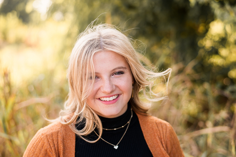 high school senior girl with blond hair wearing a rust colored sweater with hair blowing and smiling in a field in Oklahoma