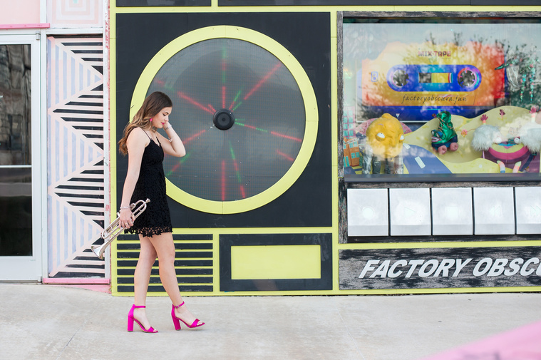 high school senior girl wearing a black dress and pink heels holding a trumpet walks parallel to camera looking down in front of Factory Obscura in Automobile Alley in Oklahoma City, Oklahoma
