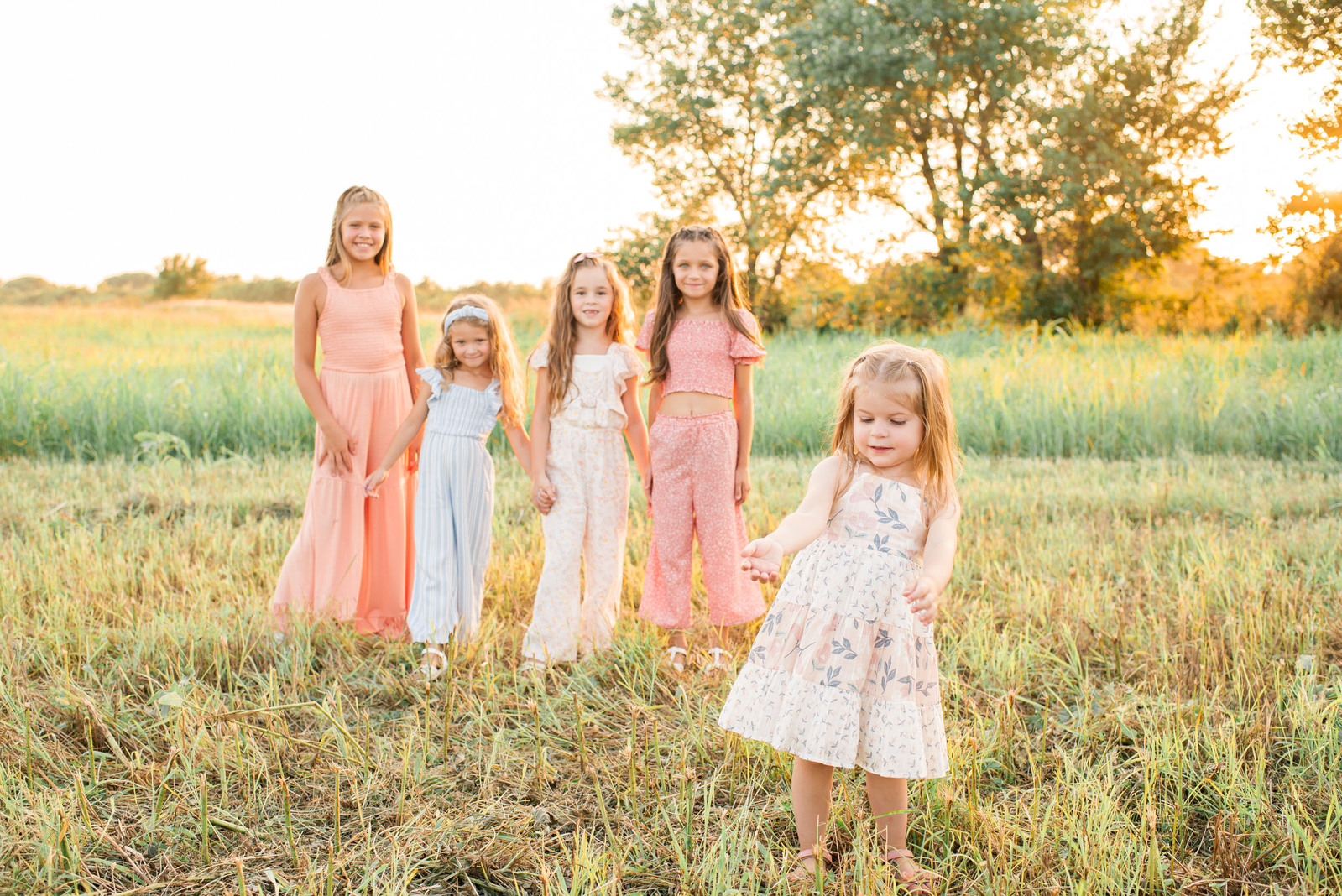 4 little girls are standing and holding hands wearing light colored rompers and a 5th toddler wearing a dress is in the foreground pointing in a field in central Oklahoma at sunset