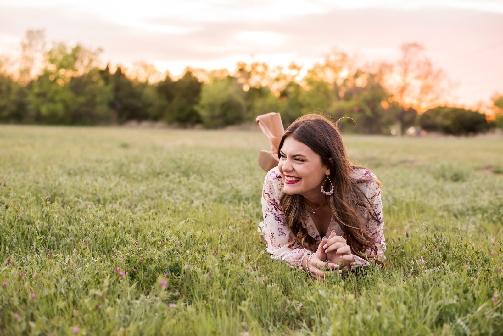 high school senior girl lays on her stomach in a green field in Oklahoma City with the sunset behind her and laughs to the side