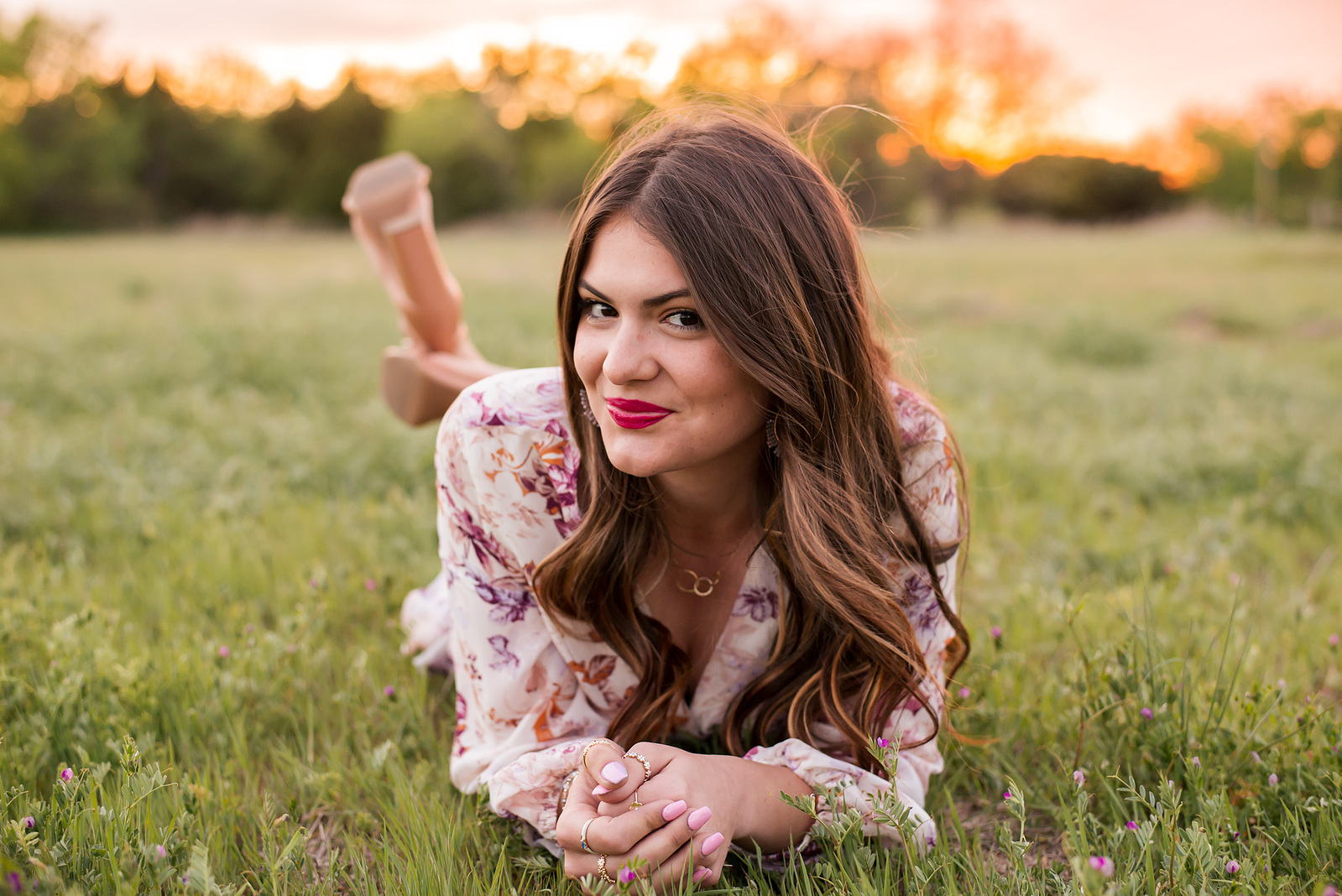 senior girl with long brown hair lays on her stomach and smiles cutely in a field in SW Oklahoma City