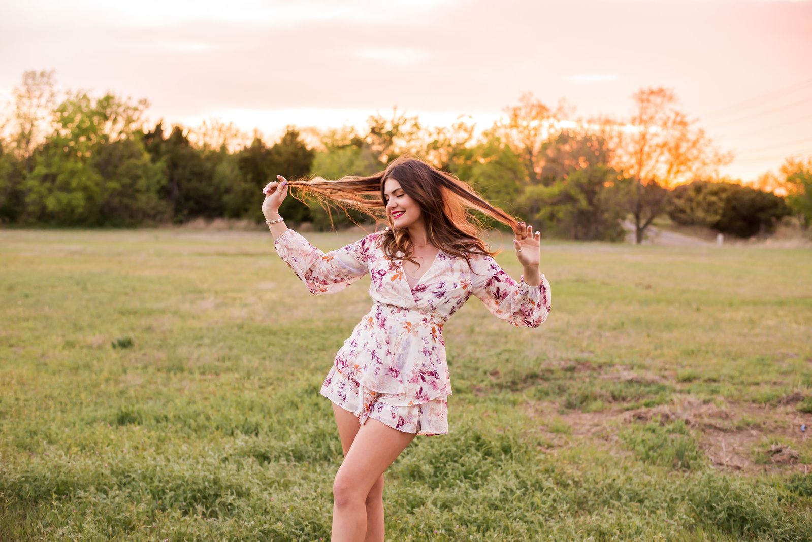 brunette senior girl wearing a floral romper stands with each hand holding out her hair and smiles in a field at sunset near Oklahoma City