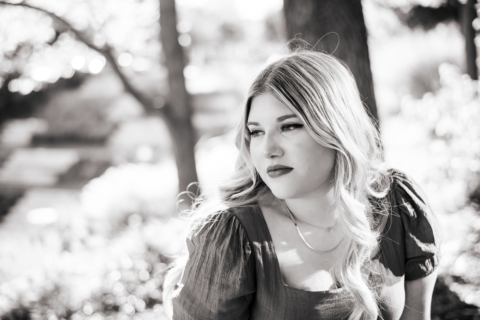 black and white photo of a senior girl looking to the side softly, sitting in front of trees and bright leaves with the sunlight coming in the side of frame at Myriad Gardens in Oklahoma City