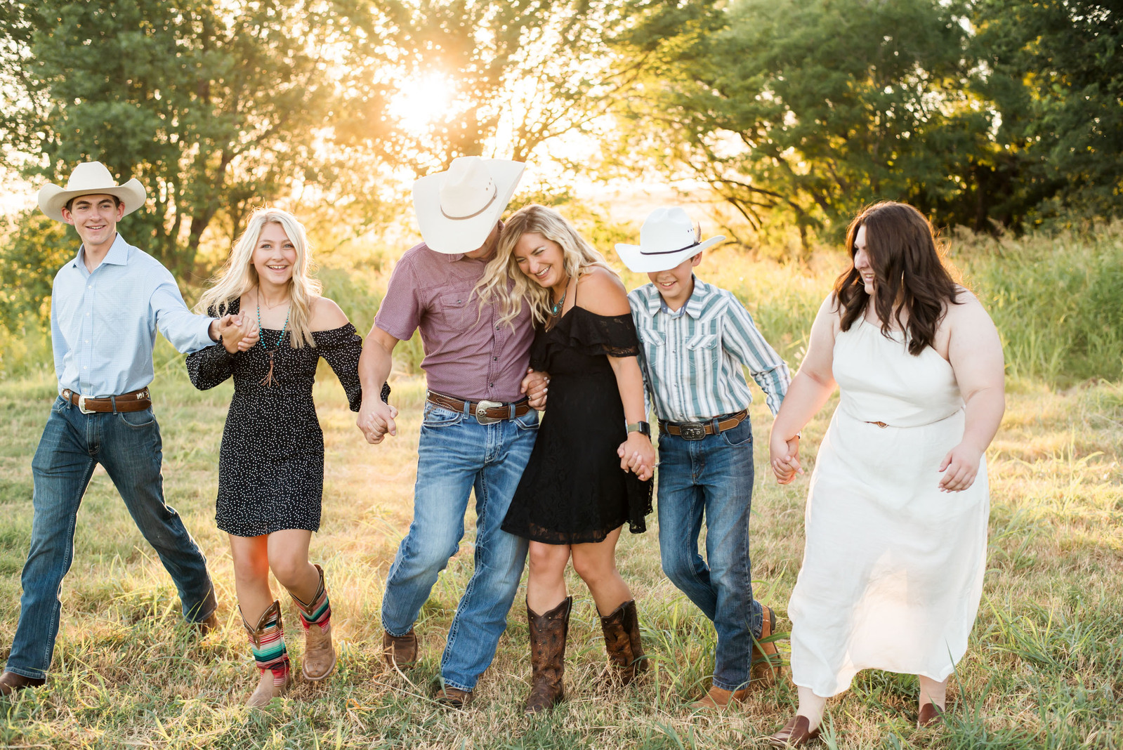 a family of 6 hold hands and bump into each other wearing western attire in a field in the country in central Oklahoma with the sun behind them