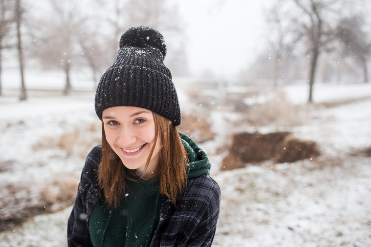 high school senior girl wearing a beanie and flannel jacket stands in Schrock Park in Tuttle, Oklahoma with snow falling around her, smiling at the camera with snow and trees in background