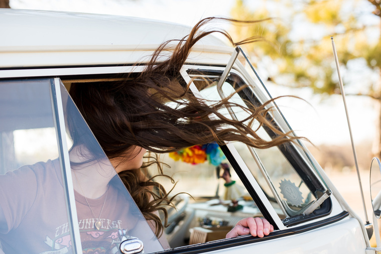 girl with long brown hair sits in the passenger side of a VW bus with the window open and her long hair flying out while she looks away