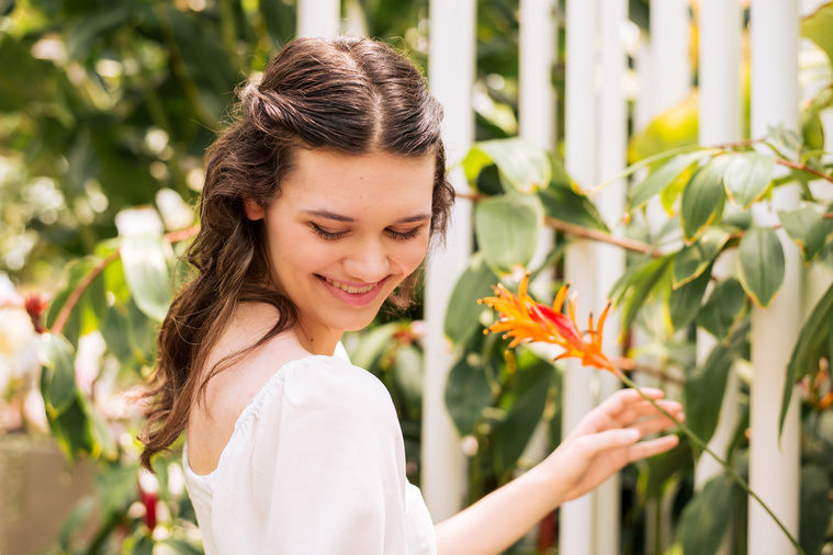 high school senior girl with long brown hair in a white blouse smiles over shoulder while holding the stem of an orange tropical flower inside the Crystal Bridge at Myriad Gardens in Oklahoma City 